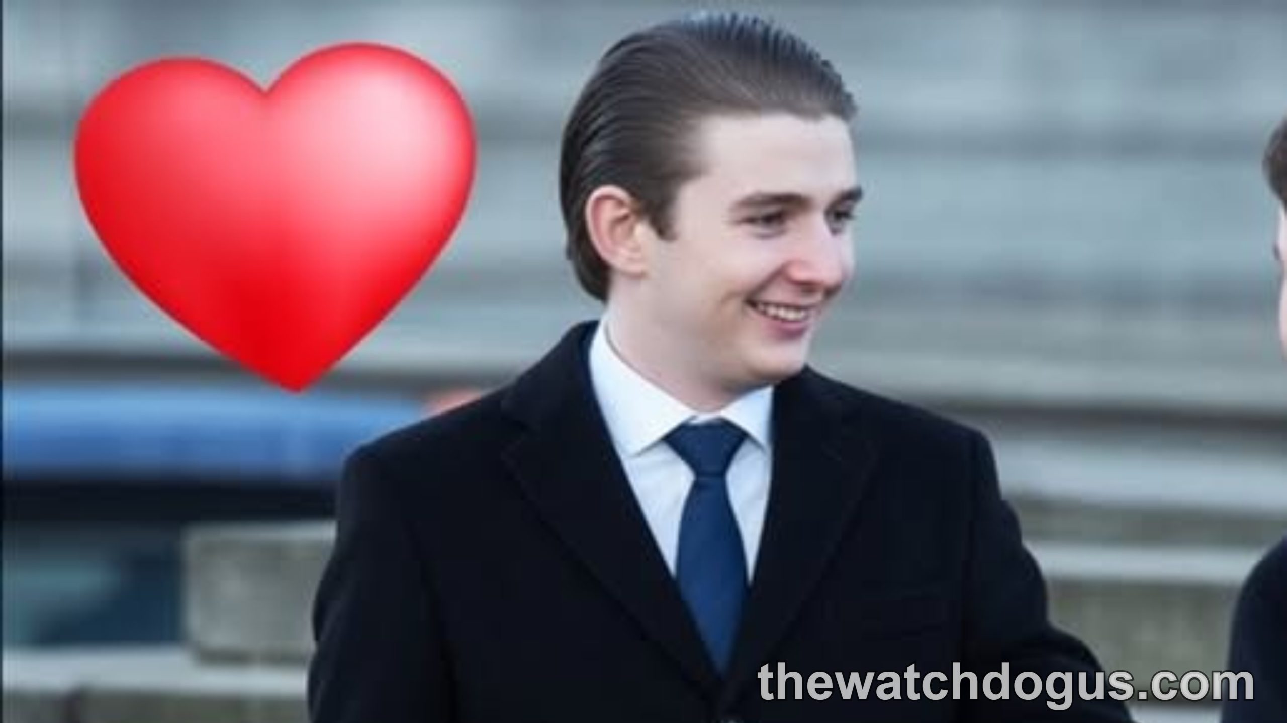 Young man in formal black suit and blue tie smiling outdoors with red heart symbol, related to Barron Trump girlfriend news