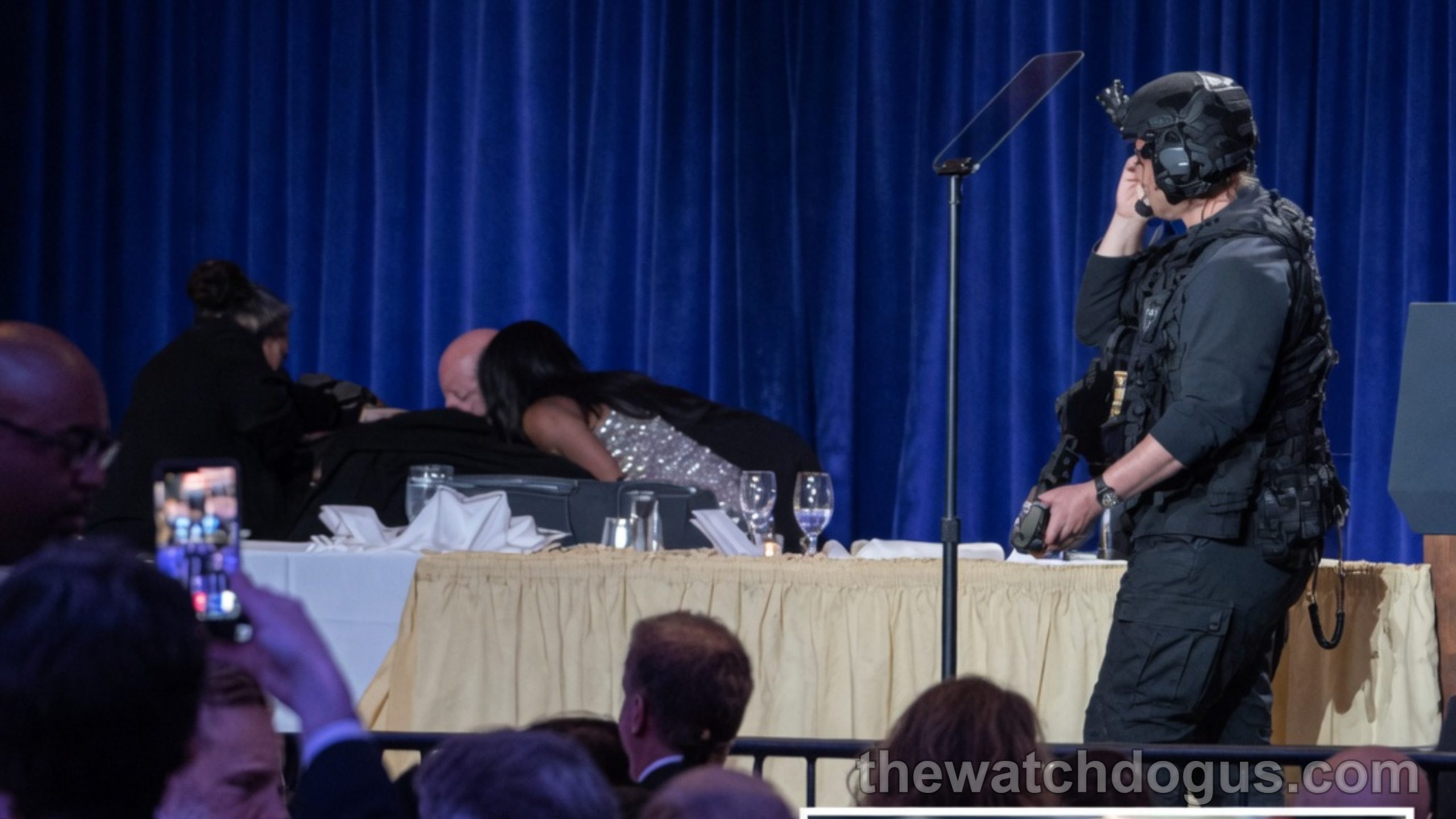 Secret Service agent in tactical gear stands at White House Correspondents dinner podium with blue curtain backdrop