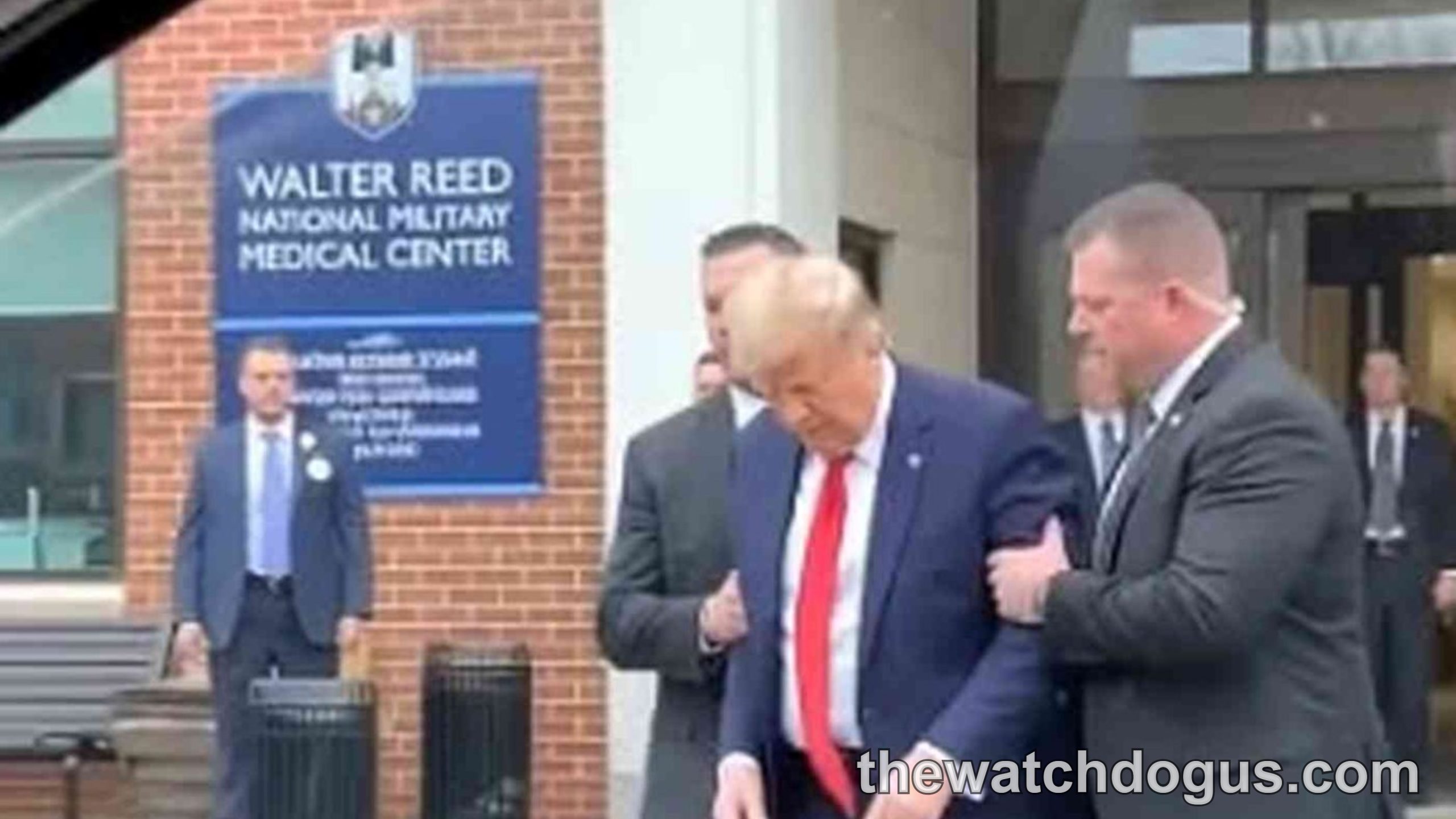 Man in dark suit and red tie walking outside Walter Reed National Military Medical Center with security personnel