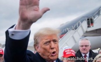 Political figure waving at rally with supporters wearing red campaign hats near airplane