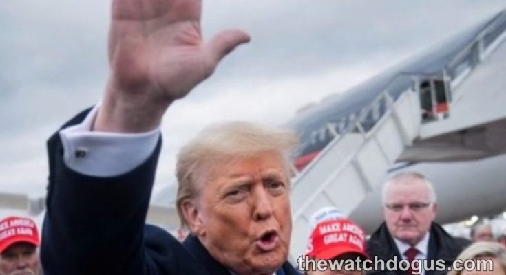 Political figure waving at rally with supporters wearing red campaign hats near airplane