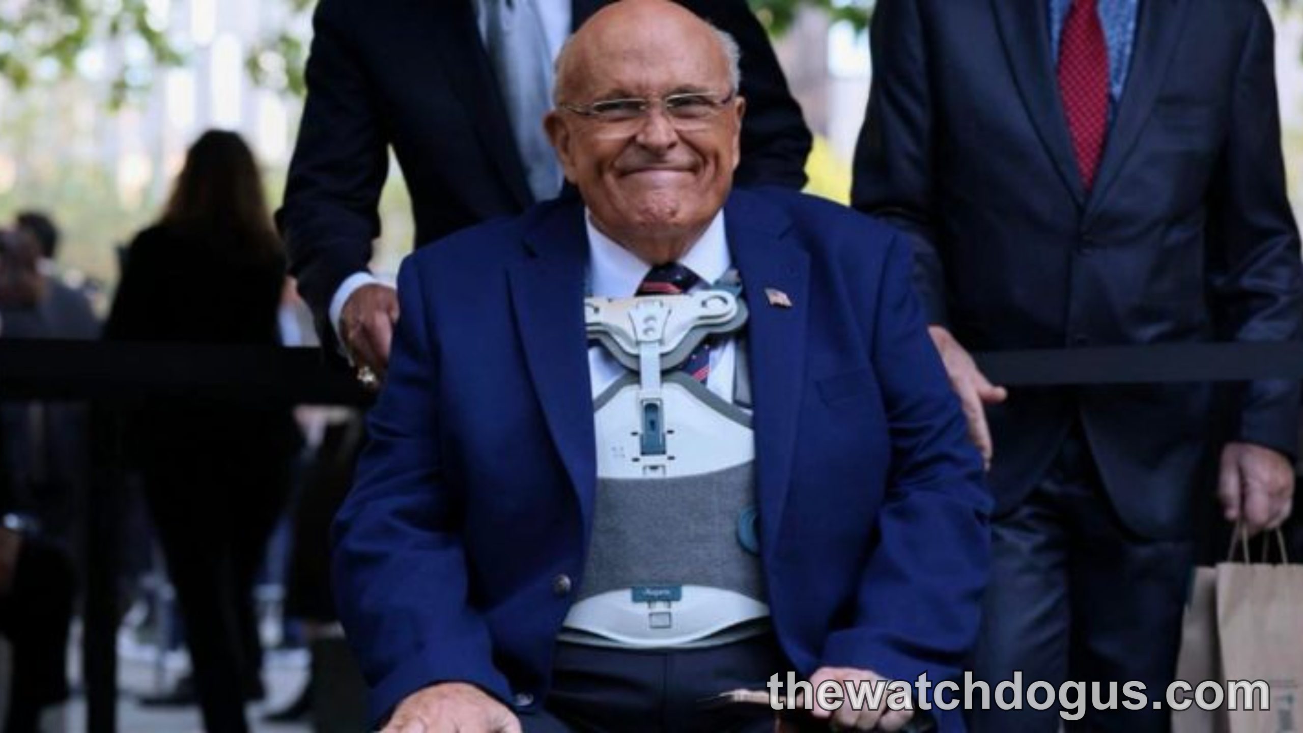 Rudy Giuliani in blue suit smiling after hospital release, accompanied by staff members outdoors